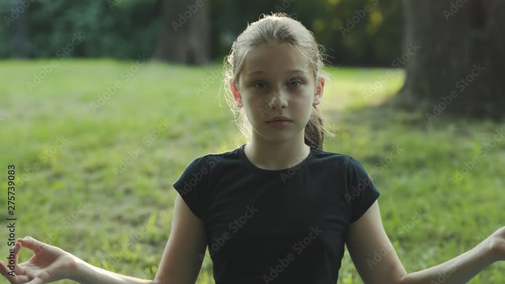 Cute serious teenage girl in black t-shirt sitting in lotus position, looking into camera. Yoga. Summertime. Park. Outdoors. City. Cute serious teenage girl in black t-shirt sitting in lotus position, looking into camera. Yoga. Summertime. Park. Outdoors. City.