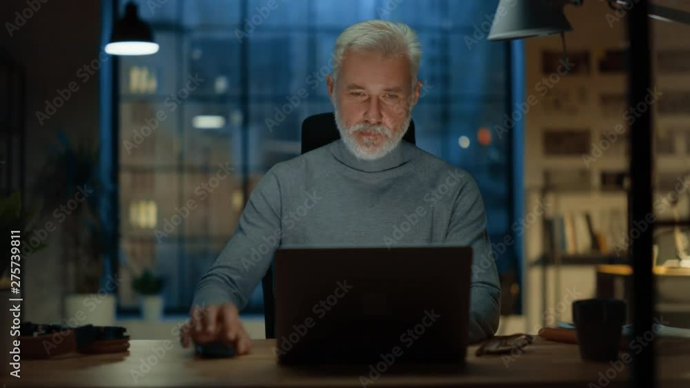 Portrait of the Handsome and Successful Middle Aged Bearded Businessman Working at His Desk Using Laptop Computer, Checks His Smartphone. Working from Cozy Home Office / Studio. Portrait of the Handsome and Successful Middle Aged Bearded Businessman Working at His Desk Using Laptop Computer, Checks His Smartphone. Working from Cozy Home Office / Studio.