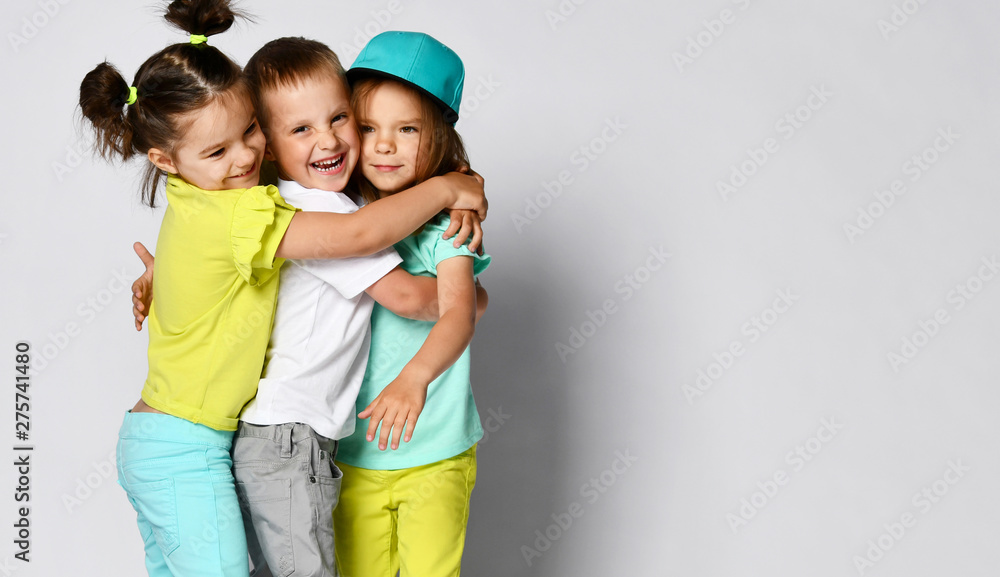 Studio portrait of children on a light background: full body shot of three children in bright clothes, two girls and one boy. Triplets, brother and sisters. hugging on camera. Family ties, friendship Studio portrait of children on a light background: full body shot of three children in bright clothes, two girls and one boy. Triplets, brother and sisters. hugging on camera. Family ties, friendship
