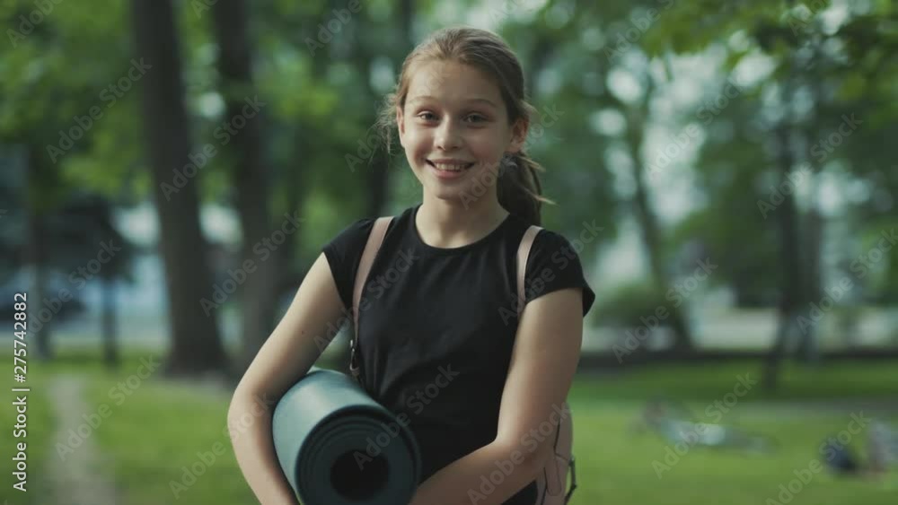 Nice Caucasian little girl with ponytail holding yoga mat in hands looking at camera. Good mood. Positive feelings. Healthy lifestyle. Outdoors. Park. Nice Caucasian little girl with ponytail holding yoga mat in hands looking at camera. Good mood. Positive feelings. Healthy lifestyle. Outdoors. Park.