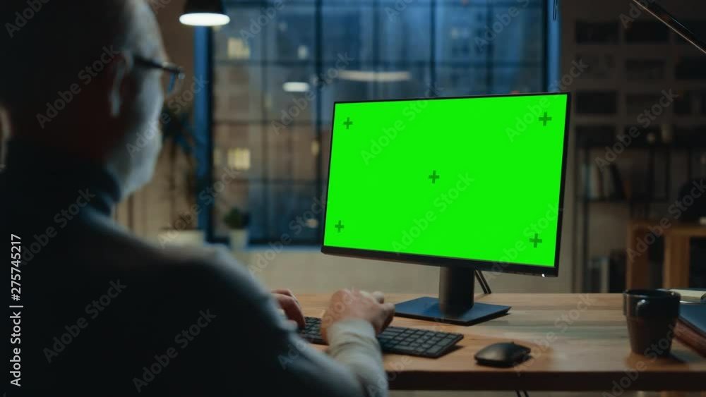 Over the Shoulder: Confident Middle Aged Man Sitting at His Desk Using Desktop Computer with Mock-up Green Screen. Evening in the Stylish Office Studio with City Window View Over the Shoulder: Confident Middle Aged Man Sitting at His Desk Using Desktop Computer with Mock-up Green Screen. Evening in the Stylish Office Studio with City Window View