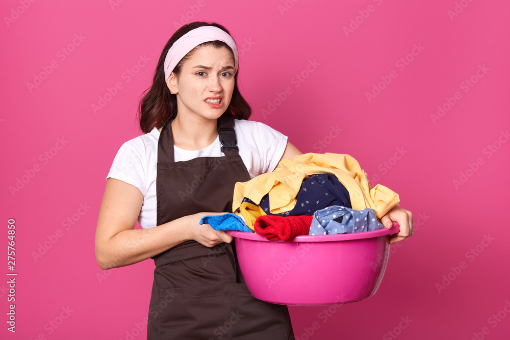 Unsatisfied upset housewife standing with unpleasant facial expression, holding pink basin full of dirty clothes, wearing light headband, white t shirt and brown apron. Household chores concept. Unsatisfied upset housewife standing with unpleasant facial expression, holding pink basin full of dirty clothes, wearing light headband, white t shirt and brown apron. Household chores concept.