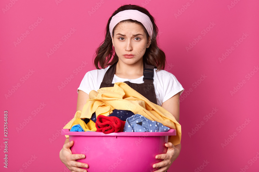 Disappointed emotional female with light headband, white t shirt and brown apron, holding laundry, exhausted from household chores, looking directly at camera. Housewife and working concept. Disappointed emotional female with light headband, white t shirt and brown apron, holding laundry, exhausted from household chores, looking directly at camera. Housewife and working concept.