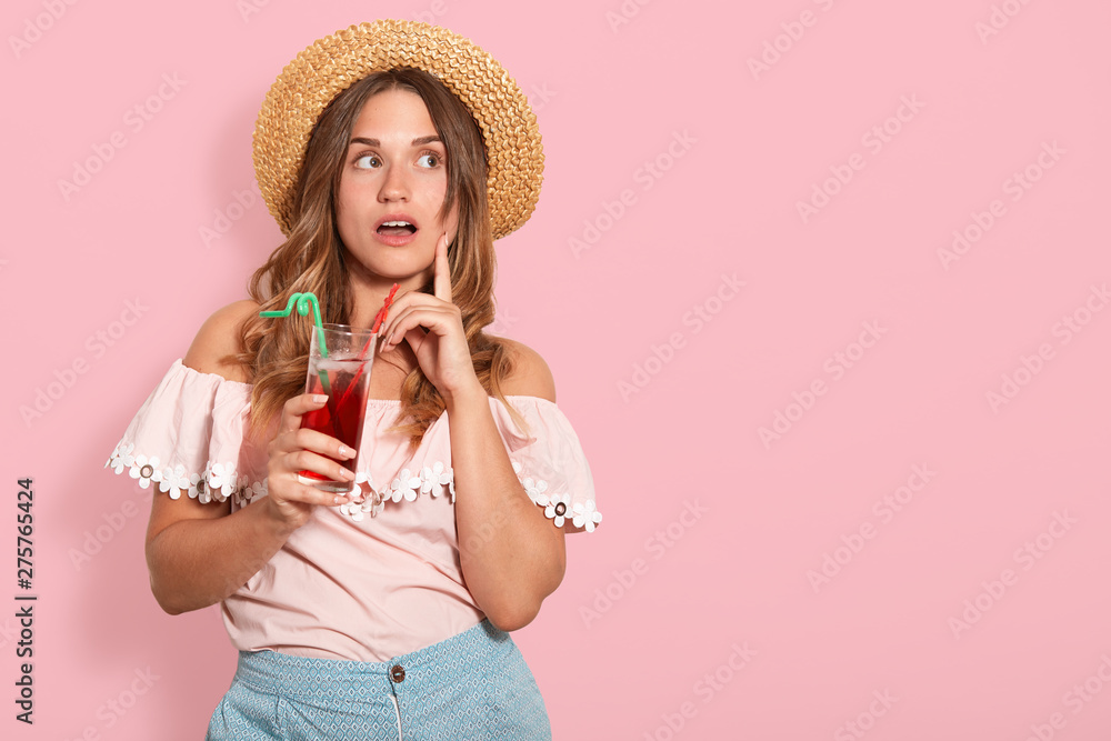 Close up studio photo portrait of thoughtful pondering female, attractive lady holding tasty beverage in hand, wearing straw sunhat and shirt, model posing isolated rose background. People concept. Close up studio photo portrait of thoughtful pondering female, attractive lady holding tasty beverage in hand, wearing straw sunhat and shirt, model posing isolated rose background. People concept.