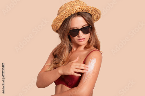 Studio shot og slim tanned girl putting sun block, redy togo to beach. Beautiful young woman enjoying sunshine on summer vacation, dressed red swimming suit and straw hat, isolated on beige background © sementsova321