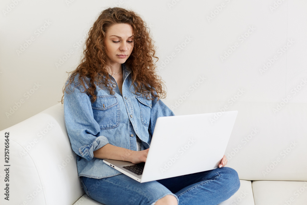Portrait of focused freelancer working online at home, using modern technology, charming female with long curly hair wearing denim jacket, sitting in cozy room on white comfortable white sofa. Portrait of focused freelancer working online at home, using modern technology, charming female with long curly hair wearing denim jacket, sitting in cozy room on white comfortable white sofa.