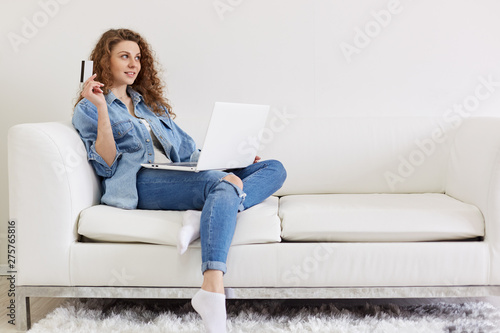 Portrait of beautiful positive lady looking aside, raising hand with credit card, making plans for online shopping, having laptop on legs, wearing jeans and jacket, having pleasant facial expression.