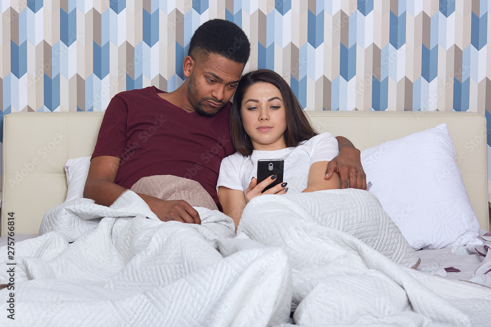 Fototapeta premium Young attractive multicultural couple in white bed, female holding smartphones in hands, wearing pajamas, searching or browsing Internet or watching film together, cheating over messages with friends.