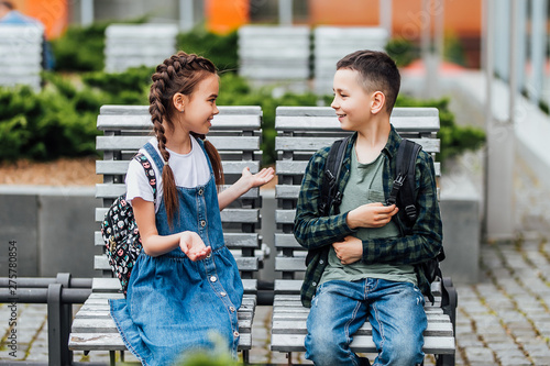 Two , little c hild with backpack sitting on the bench near the school, after finished lesson and speak with each other.