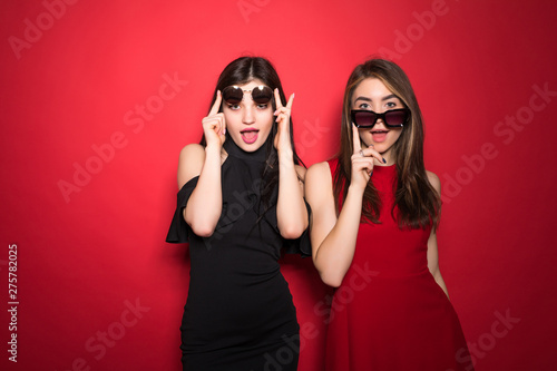 Photo of two posh women in trendy outfit and sunglasses smiling at camera isolated over red background