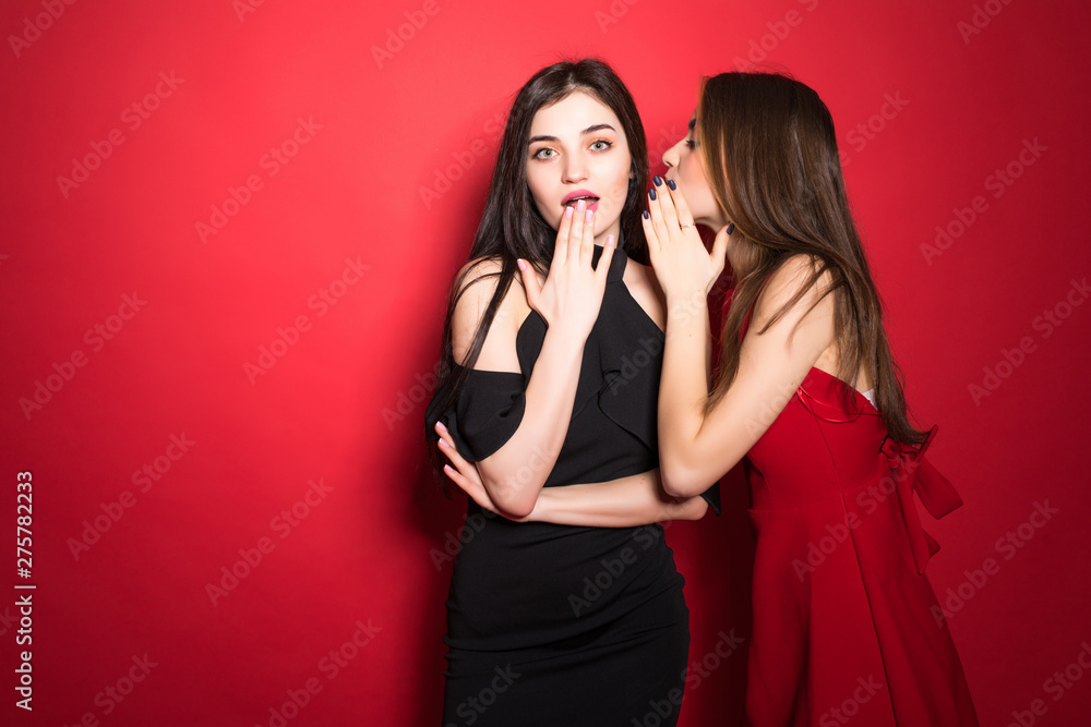 Portrait of a two cute girls dressed whispering a secret isolated over red background Portrait of a two cute girls dressed whispering a secret isolated over red background