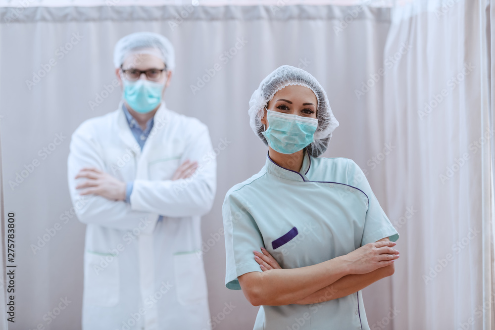Portrait of young friendly Caucasian female nurse in uniform, with mask on and hairnet standing with arms folded at clinic. In background doctor standing with arms folded, too. Portrait of young friendly Caucasian female nurse in uniform, with mask on and hairnet standing with arms folded at clinic. In background doctor standing with arms folded, too.