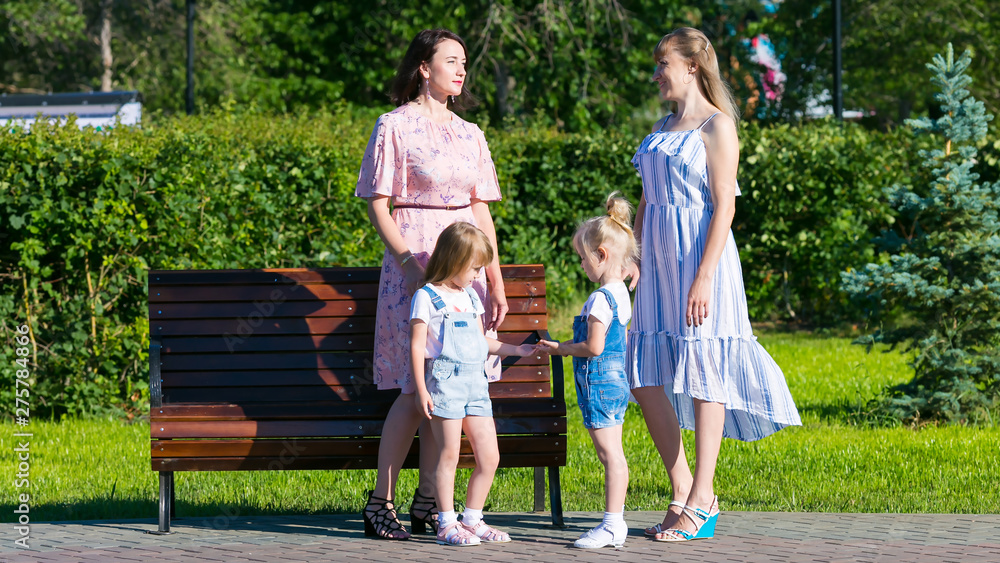 Fototapeta premium Two women with young children on the street. Women friends 35-40 years age and small daughters 4-5 years of age are talking in the park.