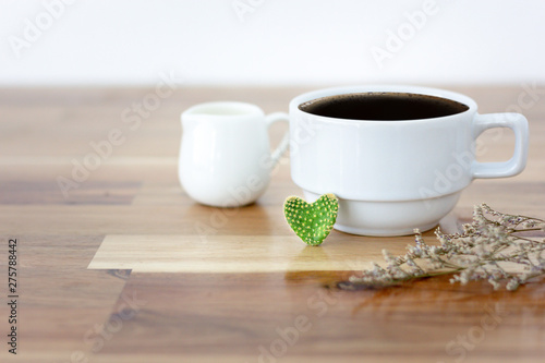 Closeup of cactus heart shape with black coffee in white cup and white cup of milk and dry leaf on wooden table. Selective and soft focus