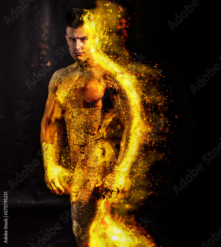 Muscular naked man covered in gold, with fire or flame on his skin, looking down sensually on light background