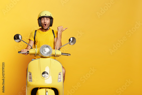 Impressed driver covers distance on yellow motorbike, wears helmet, indicates with great wonder aside, stops on road, demonstrates blank space for your advertising content, tries new vehicle.