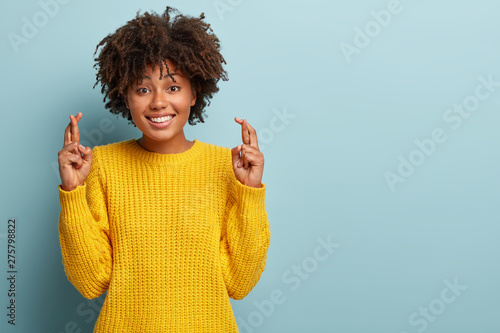 Wish and anticipation concept. Beautiful dark skinned lady stands with crossed fingers, dreams party went well, prays dream fulfill, has pleasant toothy smile, wears yellow jumper blank space on right photo