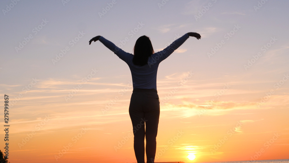 A young woman on the background of the sunset the sun on the seashore does exercises exercised by running, stretching and slumping, The girl at the sea does yoga. Portrait of: Slow motion, Sunset.