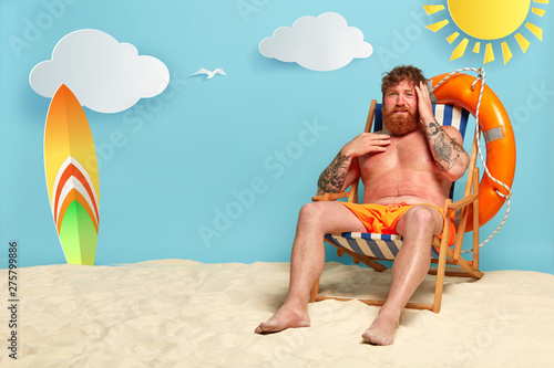 Frustrated red haired man sunbathes at sandy beach, has headache after sunstroke, wears orange shorts, sits at deck chair, has active summer rest, red sunburned skin from sun ultaviolet rays photo