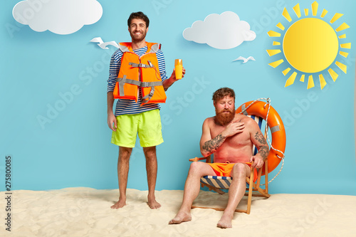 Full length shot of happy male lifeguard advertises sun screen or protection lotion, wears lifejacket, displeased friend sits near at chair, has problems with skin after sunbathing. Two men at beach photo