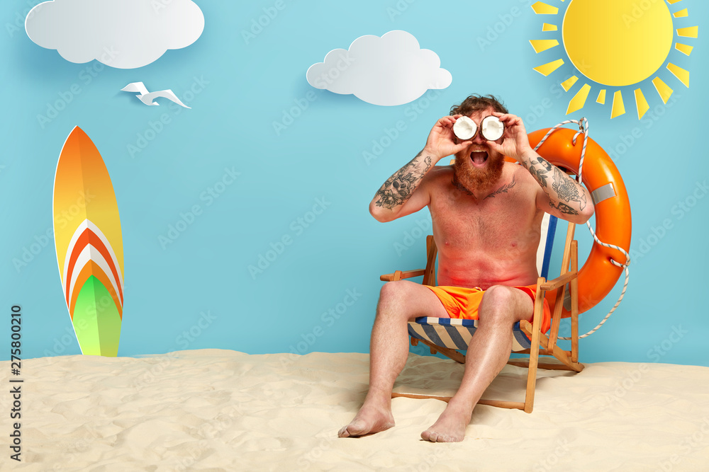 Fotografie Handsome redhead man rest at sea resort, keeps coconuts near eyes as binoculars, gazes into distance, wears shorts, sits in comfortable beach chair near lifering, surfboard