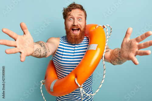 Friendly looking happy man with ginger hair and thick beard, gives hug at camera, stands with life preserver, wears striped sailor vest, says hello summer, isolated on blue. Water rescuing concept photo