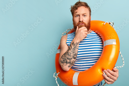 Serious redhead rescuer holds chin, has contemplative look, carries lifering, has tattooed arm, thinks abou safety swim, wears sailor vest, stands against blue wall, blank copy space. Vacation at sea photo