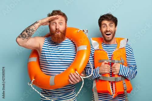 Coast security concept. Cheerful professional lifeguards pose at waterfront, wear sailor clothes, stare into distance, use safety equipment such as lifebuoy and lifejacket, support surveillance at sea