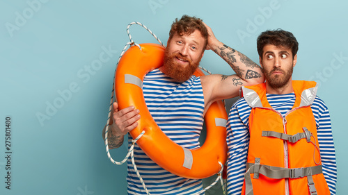 Vacation concept. Pleased red haired man tired after saving people at beach, leans at shoulder of partner who looks surprisingly aside, work together at beach, have duty to watch sea, wear life vest