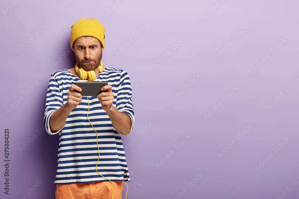 Male teenager concentrated in modern device, holds smartphone horizontally, plays online games, surfes internet during leisure time, focused into screen, wears sailor jumper, uses stereo headphones Male teenager concentrated in modern device, holds smartphone horizontally, plays online games, surfes internet during leisure time, focused into screen, wears sailor jumper, uses stereo headphones