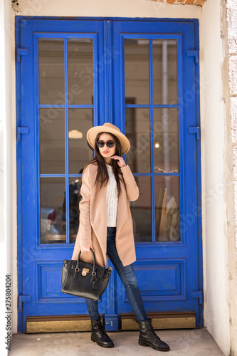 Young pretty woman woman wear in coat and hat with smile stands at the blue door on the street