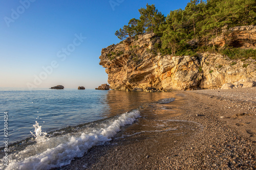 Sea bay with rocky cliff covered with growing green pine trees. Charming splashing water at seashore of empty beach in Turkey. Horizontal color photography.