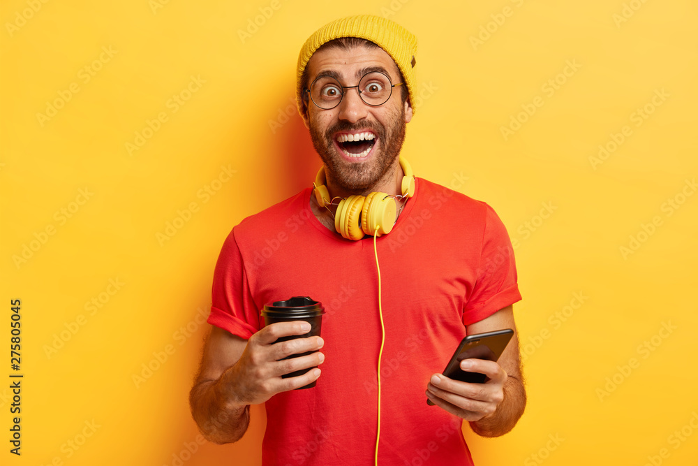 People, drinks, leisure and technology concept. Cheerful millennial guy stands with coffee cup and smartphone, has glad expression, wears red t shirt and yellow headgear, chats with friends. People, drinks, leisure and technology concept. Cheerful millennial guy stands with coffee cup and smartphone, has glad expression, wears red t shirt and yellow headgear, chats with friends.