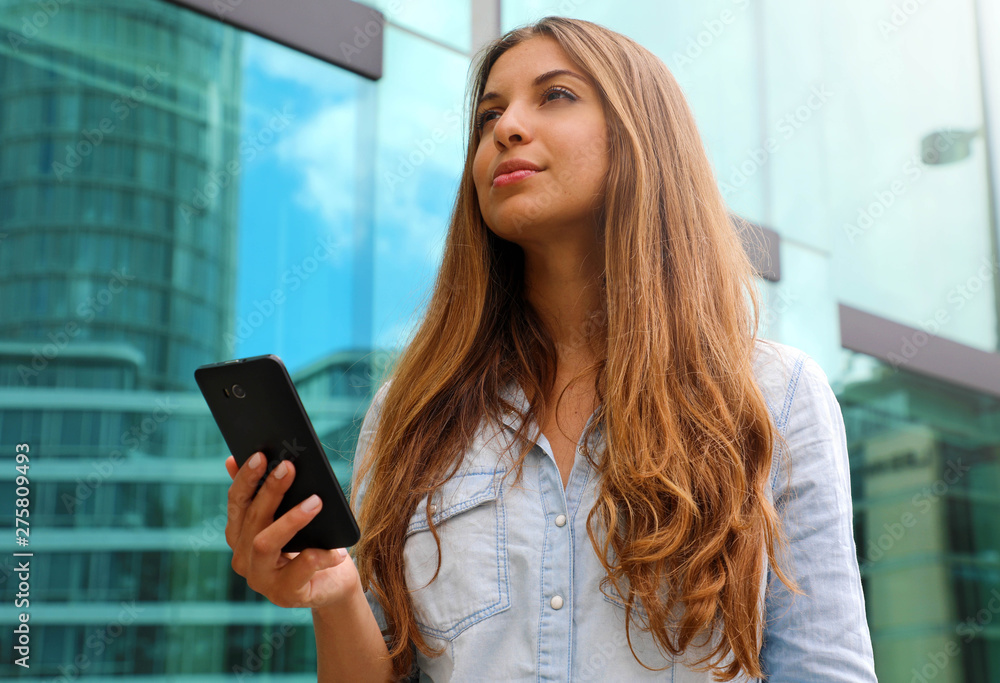 Young serious and determined business woman standing in front of modern building with phone in her hand Young serious and determined business woman standing in front of modern building with phone in her hand