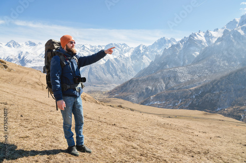 Bearded man in sunglasses and a hat with a backpack points hand at the mountains. Travel concept on the background of a mountain landscape
