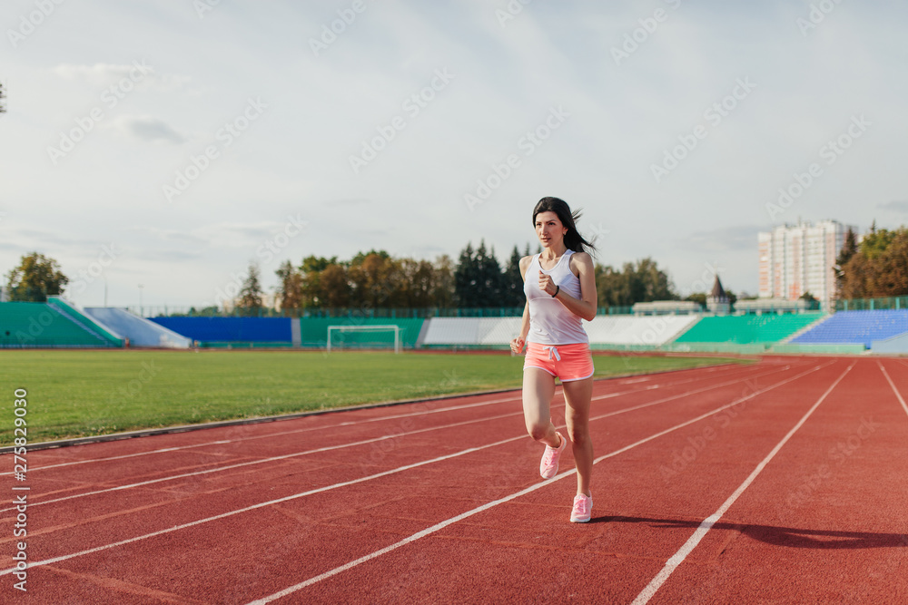 Fototapeta premium Full body of girl running track on stadium. Real front view of young woman in pink shorts and tank top and pink sneakers. Outdoors, sport