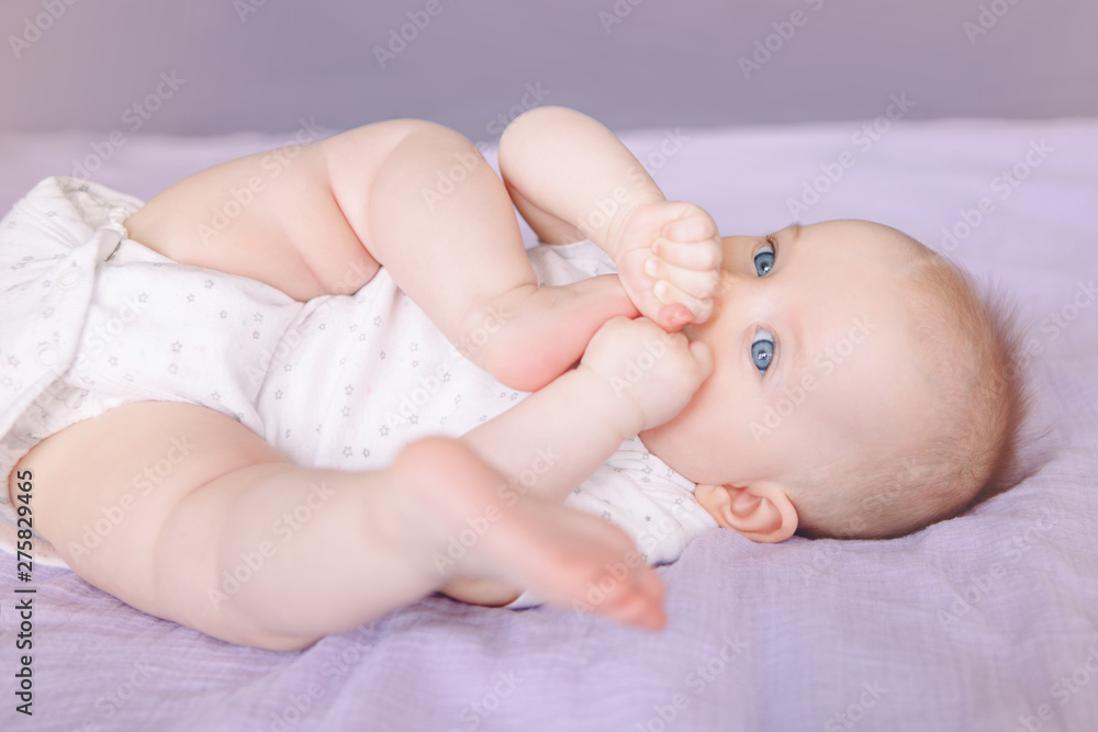 Closeup portrait of cute adorable white Caucasian infant child girl boy with blue eyes lying on bed licking sucking fingers fist. Baby teething. Happy childhood lifestyle and newborn development. Closeup portrait of cute adorable white Caucasian infant child girl boy with blue eyes lying on bed licking sucking fingers fist. Baby teething. Happy childhood lifestyle and newborn development.
