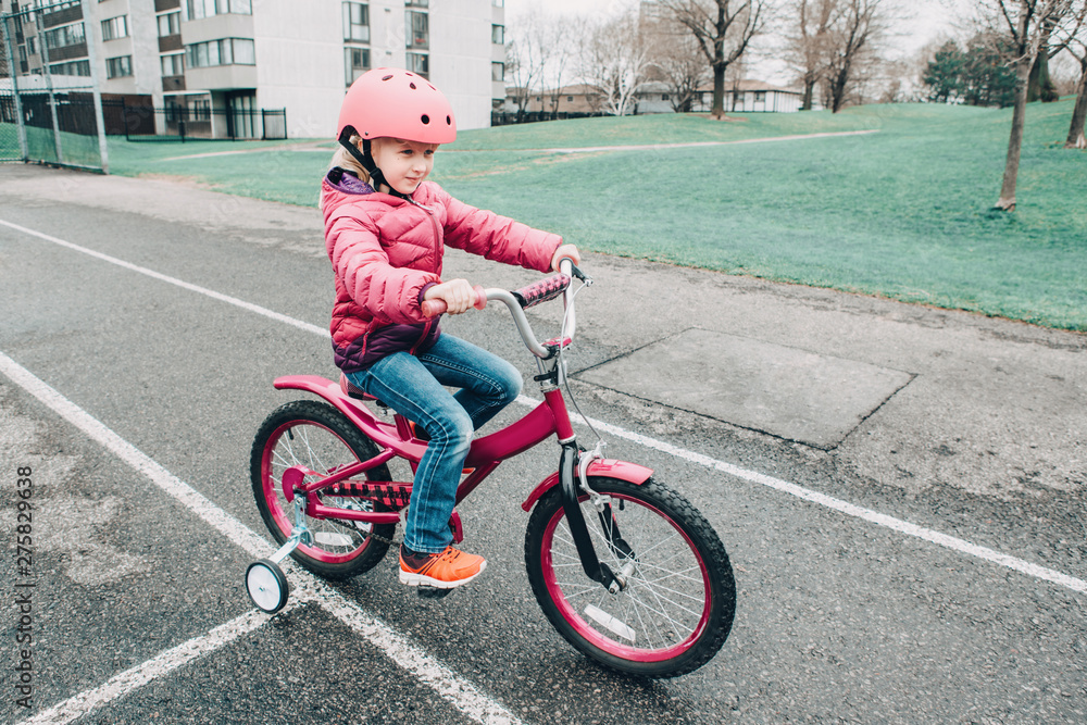 Portrait of smiling Caucasian preschooler girl riding pink bike bicycle in helmet on backyard road outside on spring autumn day. Seasonal child activity concept. Healthy childhood lifestyle Portrait of smiling Caucasian preschooler girl riding pink bike bicycle in helmet on backyard road outside on spring autumn day. Seasonal child activity concept. Healthy childhood lifestyle