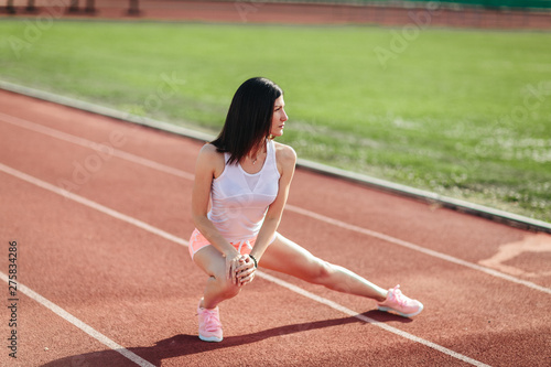 Beautiful fitness brunette girl in shorts and tank top and sneakers does a stretching exercise on the running track at the stadium outdoors. Sun shines onto her body © lena_itzy