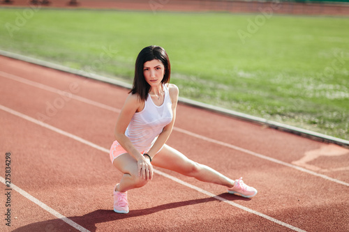 Beautiful fitness brunette girl in sportswear and sneakers does a stretching exercise on the running track at the stadium outdoors. Sun shines onto her body © lena_itzy