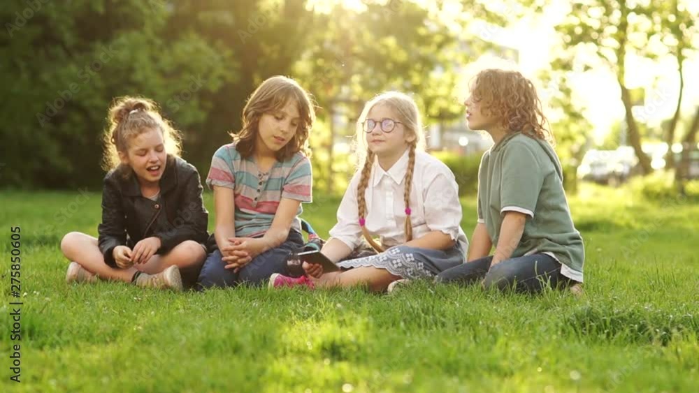 Schoolchildren boys and girls with satchels sit on the grass. Children are busy studying and discussing the smartphone. Kids and Gadgets, Back to School Schoolchildren boys and girls with satchels sit on the grass. Children are busy studying and discussing the smartphone. Kids and Gadgets, Back to School