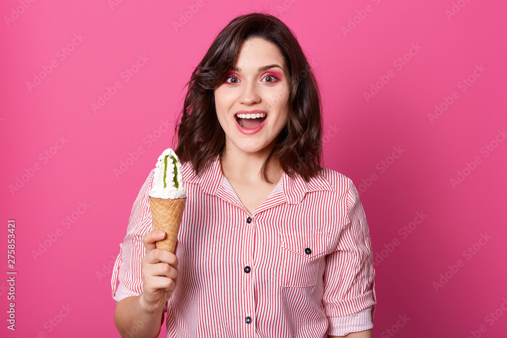 European funny young woman standing isolated over pink background in studio, opening her mouth widely with surprise, holding ice cream in one hand, being happy, wearing striped shirt. Food concept. European funny young woman standing isolated over pink background in studio, opening her mouth widely with surprise, holding ice cream in one hand, being happy, wearing striped shirt. Food concept.