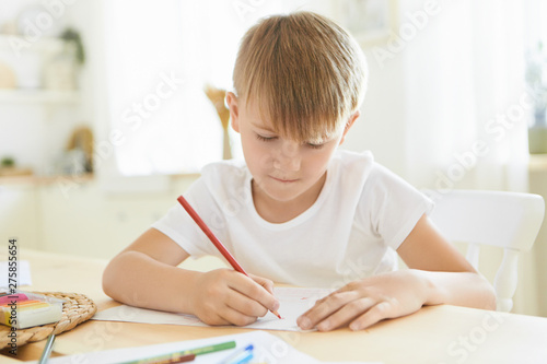 Serious focused schoolboy in white t-shirt entertaining himself indoors using red pencil drawing or sketching at wooden table isolated against stylish living room blurred interior background