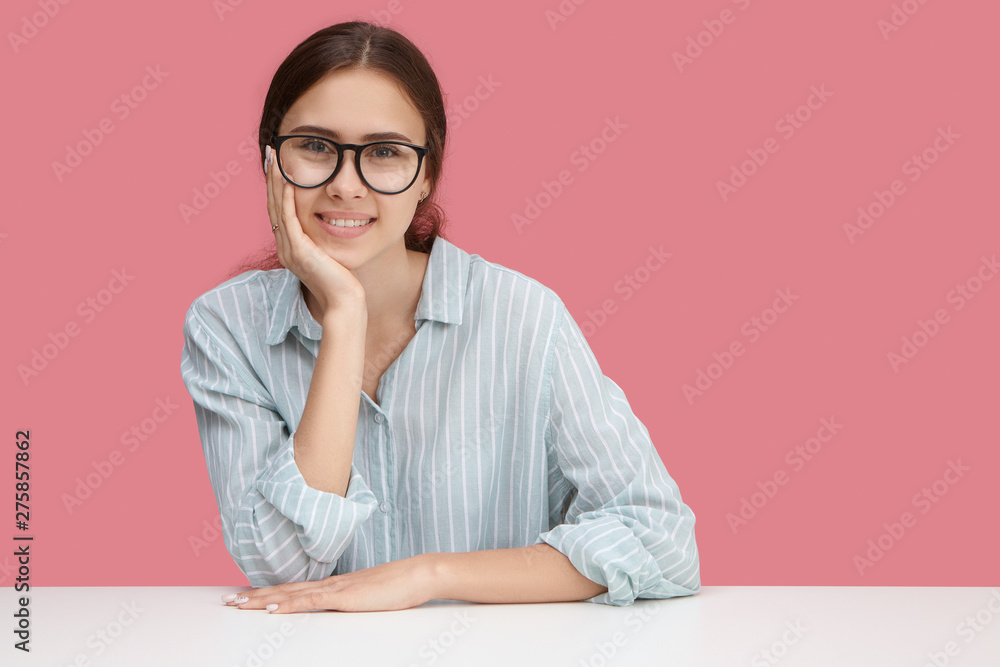 Beautiful happy young female employee wearing stylish blue shirt and spectacles enjoying working process, looking at camera with broad cheerful smile. People, lifestyle and occupation concept Beautiful happy young female employee wearing stylish blue shirt and spectacles enjoying working process, looking at camera with broad cheerful smile. People, lifestyle and occupation concept