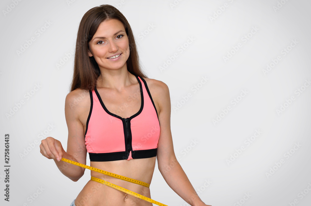 A sporty young woman in a pink top and bare belly smiles and measures her waist with a meter. A sporty young woman in a pink top and bare belly smiles and measures her waist with a meter.