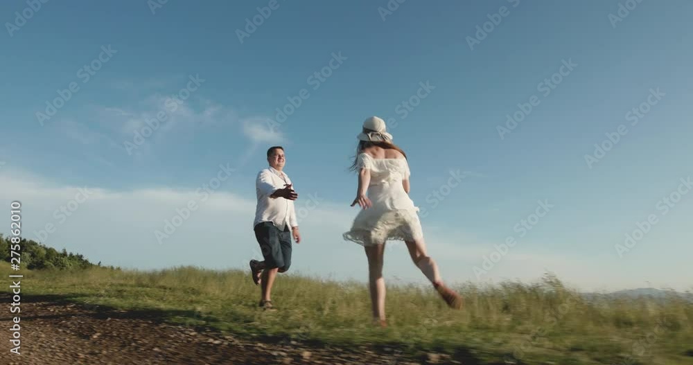 Cheerful young couple hiking in summer on mountain top. Love in the mountains. Man and woman run along the road to the horizon somewhere on the hill while sun shines over them Cheerful young couple hiking in summer on mountain top. Love in the mountains. Man and woman run along the road to the horizon somewhere on the hill while sun shines over them