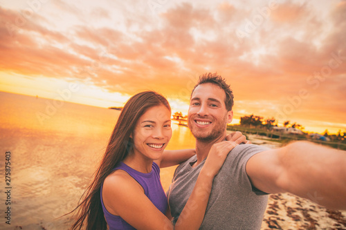 Happy selfie couple tourists on USA travel taking photo at sunset on Florida beach. Smiling Asian woman and Caucasian man, interracial relationship.