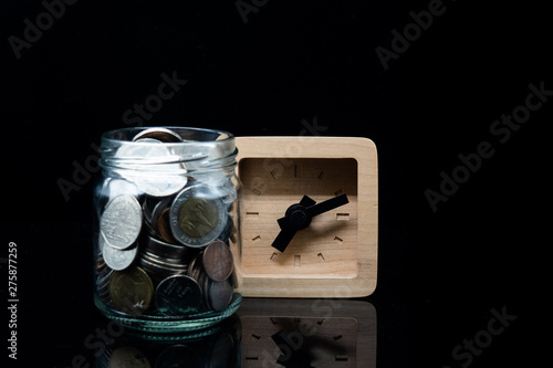 Fototapeta Naklejka Na Ścianę i Meble -  clear jar full with thai baht coin and wooden clock on black background. background concept for time factor for money saving and interest.
