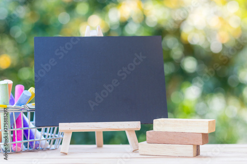 mage of empty small chalkboard on wooden easel, colorful chalk sticks in basket and blank wooden brick display label over wood table outdoor with blurred nature background. Mock-up of menu blackboard