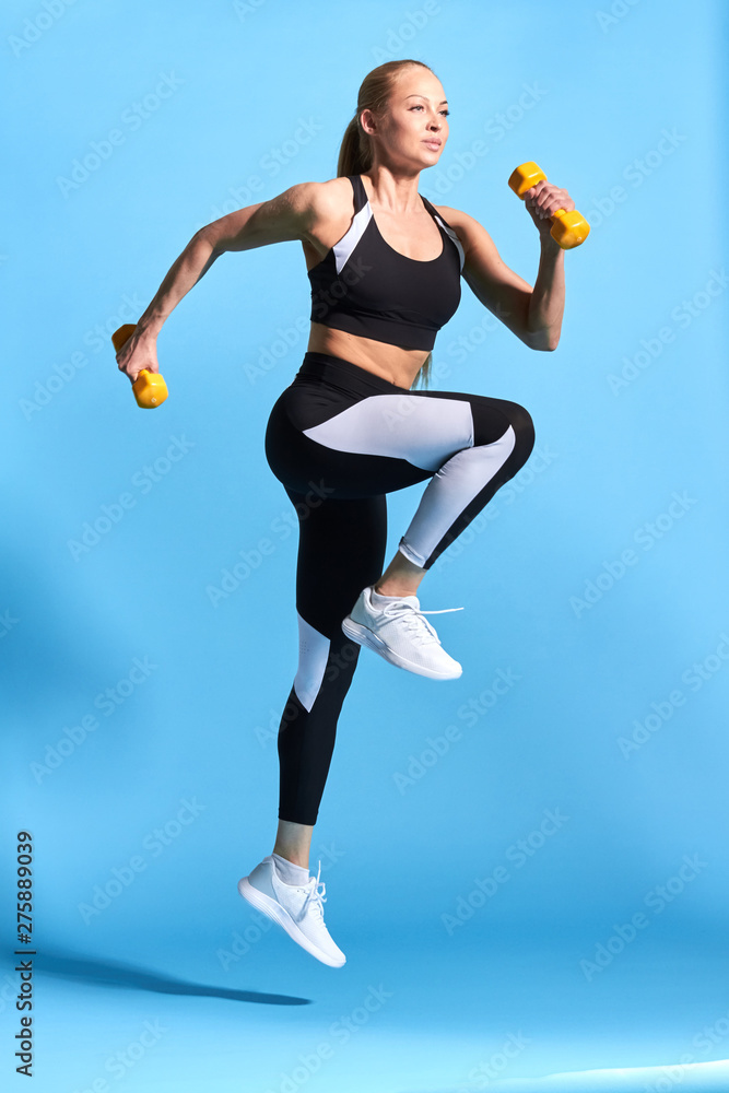 pleasant beautiful woman in stylish sportswear working out with dumbbells. full length photo. isolated blue background.motivation, long jump. body care. health care pleasant beautiful woman in stylish sportswear working out with dumbbells. full length photo. isolated blue background.motivation, long jump. body care. health care